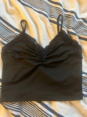 Svart linnetopp med spetsdetaljer - Snygg svart linnetopp med tunna axelband och spets längs v-ringningen. Toppen har en croppad passform och lätt rynkning framtill. Är använd men väl omhändertagen. Är tyvärr från shein med material av 100% polyester.
