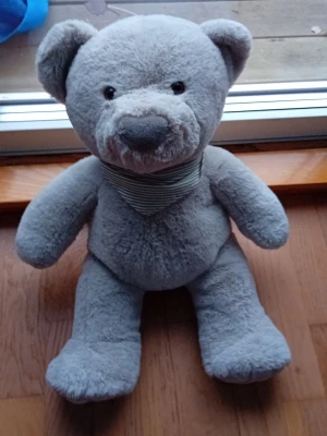 Grå mjuk nallebjörn med halsduk - Söt grå nallebjörn i mjukt plyschmaterial med svart-vitrandig halsduk runt halsen. Nallen har rund nos, små öron och broderade svarta ögon. Perfekt som gosedjur eller dekoration i rummet.