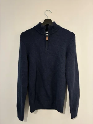 Mörkblå stickad tröja med halv dragkedja - Mörkblå stickad tröja med hög krage och halv dragkedja med läderdetalj. Tröjan har långa ärmar och ribbade muddar vid ärmslut och nederkant. Perfekt för kyliga dagar och enkel att matcha med jeans eller chinos.