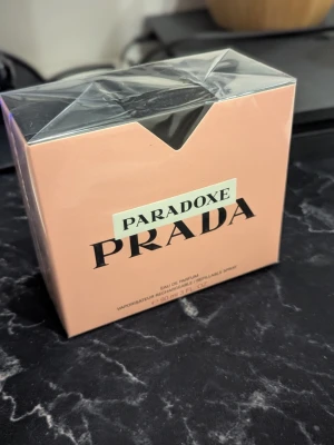 Prada Paradoxe Eau de Parfum 90ml - Prada Paradoxe Eau de Parfum i en elegant, ljusrosa kartong med svart text och stilren design. Förpackningen rymmer 90 ml och har en modern, lyxig känsla som passar perfekt för dig som gillar exklusiva dofter och snygga detaljer.