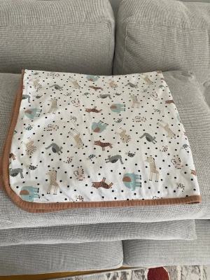 Filt med djurmönster och beige kant - Mysig filt i vitt tyg med tryck av olika djur som lejon, giraffer, zebror och elefanter i beige, brunt, blått och svart. Filtens kant är beige och tyget är mjukt, troligen bomull. Perfekt som accessoar eller till barnrummet.