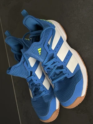 Blå Adidas sneakers med vita ränder - Snygga blå sneakers från Adidas med klassiska vita ränder på sidorna och mesh-överdel för bra ventilation. Skorna har en gummisula i ljusbrunt och detaljer i neon på plösen. Perfekta för dig som gillar sportig stil och vill sticka ut.inomhusskor 