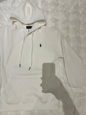 Vit hoodie från Polo Ralph Lauren - Säljer en stilren vit hoodie från Polo Ralph Lauren med klassisk huva och dragsnören. Tröjan har en broderad svart logga på bröstet och en stor magficka framtill. Tillverkad i mjukt bomullsmaterial som är skönt att ha på sig.