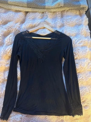 Svart långärmad spetsdetaljerad topp - Svart långärmad topp med v-ringning och fina spetsdetaljer vid halsringningen och ärmsluten. Tunn och mjuk, perfekt att styla lager på lager eller bära som den är. Enkel men ändå elegant design.