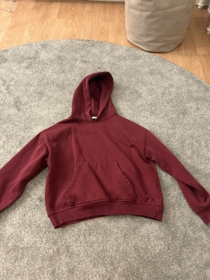 Vinröd hoodie från 157, XS - Mysig vinröd hoodie från 157 i storlek XS. Klassisk modell med huva och stor magficka framtill. Tillverkad i mjukt bomullsmaterial och har ribbade muddar vid ärmslut och nederkant. Perfekt för en avslappnad och chill stil.
