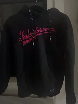 Svart Peak Performance hoodie - Svart hoodie från Peak Performance med rosa logga framtill i snyggt tryck. Klassisk modell med huva och dragsnören, samt ribbade muddar. Perfekt för en avslappnad och sportig stil. Materialet är mjukt och bekvämt. 