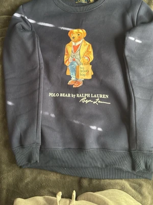 Mörkblå Polo Bear sweatshirt Ralph Lauren - Mörkblå sweatshirt från Ralph Lauren med ikoniska Polo Bear-trycket framtill. Björnen har en beige kappa, jeans och håller en 'big brown bag'. Tröjan har rund hals, ribbade muddar och är gjord i mjukt bomullsmaterial. Perfekt för en avslappnad och trendig stil. Använts 1 gång