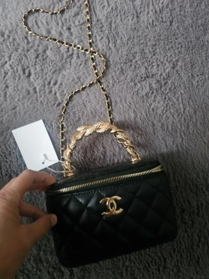 Svart quiltad handväska l - Snygg svart quiltad handväska med guldfärgad kedja och flätat handtag. Väskan har ett klassiskt CC-spänne framtill och dragkedja upptill. Insidan är fodrad i rött och har en liten spegel. Perfekt för dig som gillar lyxiga detaljer.