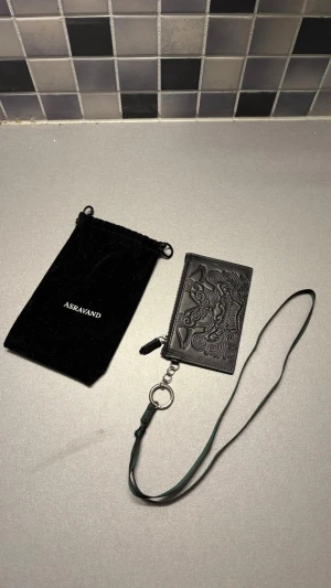 Svart korthållare från ASRAVAND - Snygg svart korthållare i läder från ASRAVAND med präglat mönster och logga. Har flera kortfack, dragkedjeförsett myntfack och en avtagbar rem med metallring. Perfekt för att hålla koll på kort och småsaker.