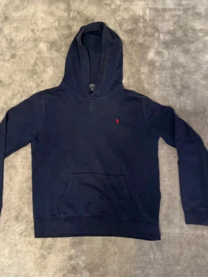 Mörkblå hoodie från Polo Ralph Lauren - Snygg mörkblå hoodie från Polo Ralph Lauren med klassisk huva och känguruficka. Liten röd broderad logga på bröstet. Perfekt för en avslappnad och stilren look. Tillverkad i mjukt bomullsmaterial som känns skönt mot huden. Storlek 170A/XS