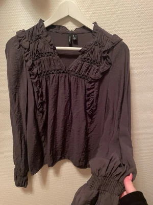 Mörkgrön blus med volang  -  Mörkgrön blus med volangdetaljer och smockade muddar vid ärmsluten. Blusen har v-ringning och dekorativa sömmar framtill. 