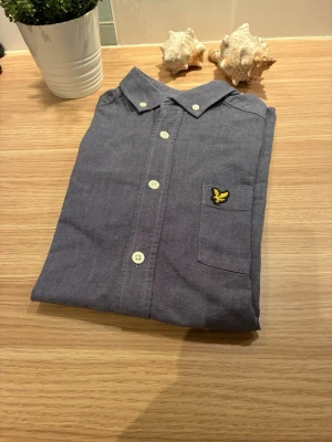 Blå skjorta från Lyle & Scott - Klassisk blå skjorta från Lyle & Scott med vit knappstängning och bröstficka med den ikoniska gula loggan. Skjortan har lång ärm och button down-krage. Perfekt för en stilren och avslappnad look.