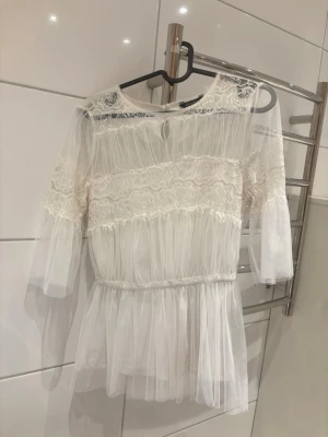 Vit spetsblus med volanger - Säljer en superfin vit blus i transparent mesh med spetsdetaljer och volanger. Blusen har trekvartsärmar, rund halsringning och resår i midjan för en snygg siluett. Perfekt för dig som gillar romantisk och trendig stil.