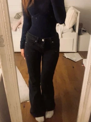Svarta flared jeans med hög midja - Säljer ett par svarta jeans med hög midja och flared ben. Jeansen har klassiska fem fickor, bälteshällor och silverfärgade knappar. Materialet ser ut att vara stretchigt denim för en bekväm passform. Perfekta till en trendig vardagslook.