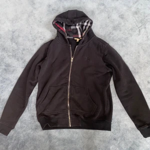 Svart zip-hoodie från Burberry  - Svart hoodie från Burberry i bomull med klassiskt rutigt foder i huvan. Tröjan har dragkedja framtill, två fickor och broderad logga på bröstet. Perfekt för dig som gillar stilrena och exklusiva plagg.  Storlek M. 