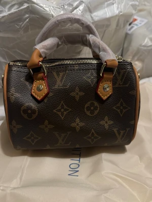 Monogram väska - Säljer en handväska i klassiskt brunt monogramcanvas med beige läderdetaljer och guldfärgade metalldelar. Väskan har två handtag och dragkedja upptill. Perfekt för dig som gillar lyxiga accessoarer och ikonisk design.