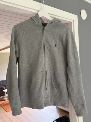 Grå zip hoodie från Polo Ralph Lauren - Klassisk grå hoodie med dragkedja från Polo Ralph Lauren. Tröjan har huva, två fickor framtill och den ikoniska lilla loggan broderad på bröstet. Tillverkad i mjukt bomullsmaterial som är skönt att bära. Perfekt för en avslappnad och stilren look. Skulle säga att tröjan passar 160-175