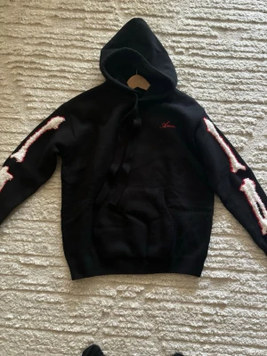Svart hoodie med skelettärmar - Svart hoodie från Amiri med huva och stora snören. På båda ärmarna finns vita och röda skelettmotiv som ger en edgy vibe. Liten röd Amiri-logga på bröstet och klassisk magficka framtill. Mjuk och skön bomullskänsla.