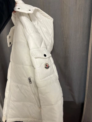 Vit dunjacka från Moncler med huva - Snygg vit dunjacka från Moncler med huva och Moncler-logga på ärmen. Jackan har en ficka med knapp på ärmen och en snedställd dragkedjeficka på sidan. Perfekt för kalla vinterdagar och riktigt stilren look. Äkta!! Skriv för fler bilder