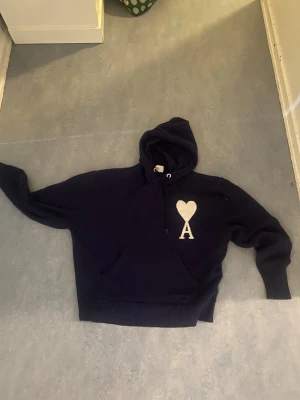 Mörkblå hoodie från Ami Paris - Snygg mörkblå hoodie från Ami Paris med stor vit hjärtlogga och bokstaven A på bröstet. Klassisk känguruficka framtill och justerbar huva med dragsko. Tillverkad i mjukt material som känns skönt mot huden. Perfekt för en avslappnad och trendig look.
