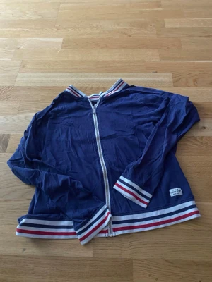 Blå vindjacka från lager 157 - Snygg blå vindjacka från lager 157 med vita och röda ränder på muddar, krage och nederkant. Jackan har dragkedja framtill och en sportig look som passar perfekt till vardagsstilen. Tillverkad i ett lätt material.