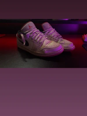 Nike Air Jordan 1 Low Vit/Svart/Röd - Säljer ett par Nike Air Jordan 1 Low sneakers i vitt läder med svarta swoosh-detaljer och röda inslag på hälen och loggan. Klassisk siluett med perforerad tåbox och vita skosnören. Perfekta för dig som gillar streetwear och ikoniska sneakers.
