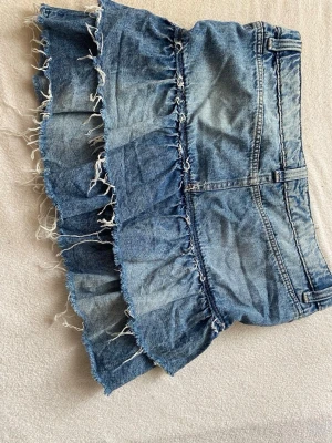 Blå volangkjol i denim med fransar - Säljer en blå jeanskjol med volanger och råa fransade kanter. Kjolen har klassisk femficksmodell, dragkedja och knapp framtill. Perfekt för dig som gillar en avslappnad och trendig stil.