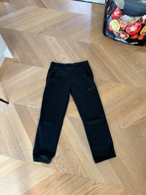 Svarta mjukisbyxor från Nike - Säljer ett par svarta mjukisbyxor från Nike med svart logga på benet. Byxorna har snören i midjan och två sidofickor. Tillverkade i ett mjukt och bekvämt material som passar perfekt för chill eller träning. Klassisk rak passform och stilren design. Lite liten i storleken så skulle säga att den passar mer åt S håller funkar möjligtvis på M. Säljer pga för små. ALDRIG andvända, prislapparna finns kvar 