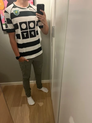 Randig fotbollströja Sporting Lissabon - Säljer en grönvit randig fotbollströja från Sporting CP med klubbmärke på bröstet och sponsortryck framtill. Tröjan har korta ärmar och rund hals. Perfekt för dig som vill ha en klassisk supportertröja till match eller träning. Trycket har lossnat där bak. Modellen är 179
