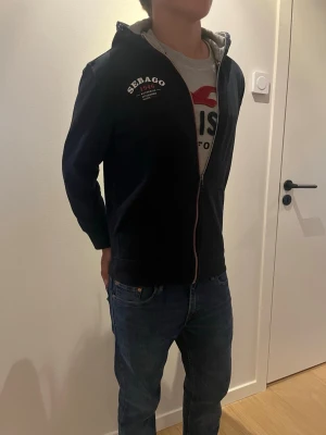 Marinblå Sebago hoodie med dragkedja - Marinblå hoodie från Sebago med vit logga på bröstet och dragkedja framtill. Modellen har huva med grått innerfoder och ribbade muddar. Perfekt för en avslappnad och sportig stil. Den är liten sliten på ett av snörena. Modellen är 179