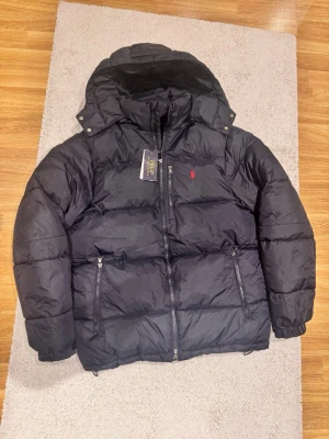 Marinblå ralph lauren puffer jacka - Marinblå pufferjacka från Polo Ralph Lauren med huva och dragkedja framtill. Jackan har två framfickor med dragkedja och den klassiska röda loggan broderad på bröstet. Perfekt för kalla vinterdagar och riktigt stilren look.