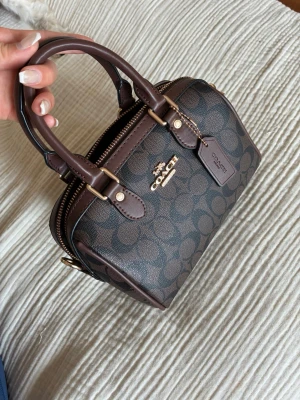 Brun handväska från Coach - Snygg brun handväska från Coach med klassiskt monogrammönster och guldfärgade detaljer. Väskan har två handtag, dragkedja upptill och en tag med Coach-logga. Tillverkad i tåligt konstläder med mörkbruna detaljer. Perfekt för dig som gillar stilrena accessoarer. Insidan har någora fläckar men inget som stör och i övrigt näst intill perfekt skick 🥰