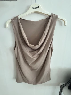 Beige topp - Säljer en snygg beige linnetopp lite likt en off shoulder topp. Toppen är ärmlös och har en mjuk, följsam passform som ger en avslappnad men ändå stilren look. Perfekt att matcha med jeans eller kjol för en trendig outfit.