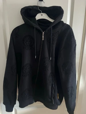 Svart hoodie med dragkedja - Säljer en svart hoodie med dragkedja framtill och huva med snören. Hoodien har ett ton-i-ton mönster. Tillverkad i mjukt material och har ribbade muddar vid ärmar och nederkant.