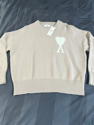 Ami tröja - Snygg beige stickad tröja med vit hjärtdetalj och bokstaven A på bröstet. Tröjan har rund halsringning, ribbade muddar och nederkant. Perfekt för dig som gillar stilrena plagg med en twist.