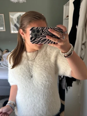 Vit fluffig topp från WERA - Supermjuk och fluffig vit topp från WERA med korta ärmar och rund hals. Toppen har en oversized känsla och är perfekt för dig som gillar en mysig och trendig stil. Materialet är syntet och ger en cool, lurvig look.