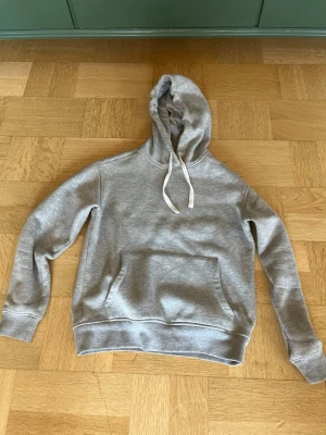 Grå hoodie med biltryck på ryggen - Snygg grå hoodie med huva och vita dragsnören. Stor känguruficka framtill och ett stort svartvitt tryck med racingbil och texten 'MAGGIORE' på ryggen. Mjuk bomullskänsla och avslappnad passform, perfekt för en cool streetstil.