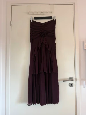 Vinröd/lila långkjol - Säljer en vinröd/lila långkjol med volangdetaljer och en dekorativ rosett framtill. Kjolen har en romantisk look med mjukt, flytande tyg och passar perfekt till speciella tillfällen. Stilren och elegant design. Den är helt ny, aldrig använd