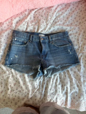 Blå jeansshorts med fransig kant - Säljer ett par klassiska blå jeansshorts med fransig kant och slitna detaljer. Shortsen har fem fickor, bälteshällor och knappgylf. Perfekta för varma dagar och en avslappnad stil. Använda 1 gång