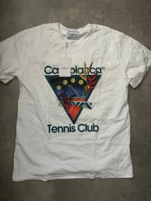 Vit Casablanca Tennis Club t-shirt - Vit t-shirt från Casablanca med färgglatt tryck framtill som visar tennisbollar, växter och texten 'Tennis Club'. Klassisk rund hals och korta ärmar. Perfekt för dig som gillar sportig och trendig stil.