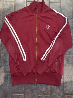 Vinröd Fred Perry ziptröja - Säljer en vinröd ziptröja från Fred Perry med vita ränder längs ärmarna och klassisk logga på bröstet. Tröjan har hög krage, muddar vid ärmslut och nederkant samt dragkedja framtill. Perfekt för en sportig och retro look.