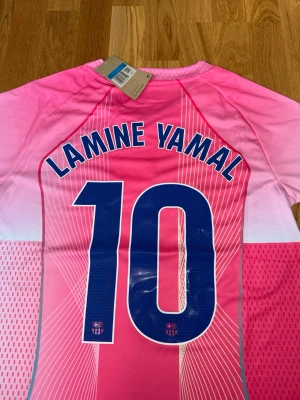 Barça rosa matchtröja Lamine Yamal - Säljer en Barcelona matchtröja i rosa och vitt med Lamine Yamal och nummer 10 på ryggen. Tröjan har Nike-logga, Barça-emblem och Spotify som sponsor framtill. Tillverkad i lätt, ventilerande funktionsmaterial och har korta ärmar.
