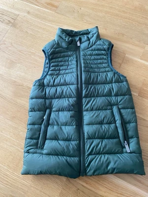Grön dunväst från 157 Funk - Snygg grön dunväst från 157 Funk med hög krage och dragkedja framtill. Västen har två fickor med dragkedja och quiltad design som ger en sportig look. Perfekt att slänga över en hoodie eller tröja för extra värme.