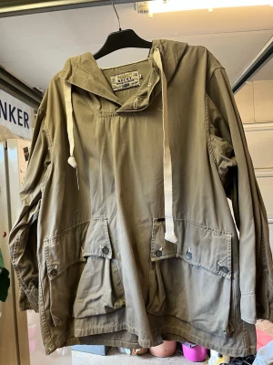 Retro/Vintage Olivgrön anorak med huva från HOLY - Säljer retro/vintage olivgrön anorak från ROCKY med stor huva och vita dragsnören. Jackan har två stora fickor framtill med knappar och en avslappnad passform. Tillverkad i slitstarkt bomullstyg, perfekt för lager på lager och streetwear. Axel till axel 55cm. Armhåla till armhåla 60cm. Axel till midja 60cm. Midja: 75cm. 
