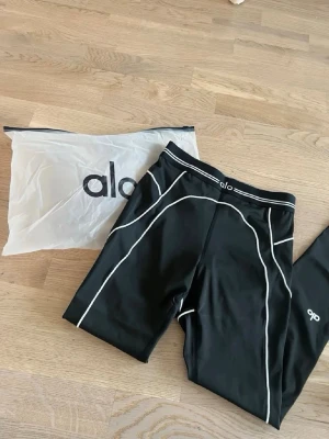 Svarta tights från Alo med vita sömmar - Snygga svarta träningsleggings från Alo Yoga. Kommer med originalpåse. Säljes pga de är för små i storleken. Prislapp finns kvar. 