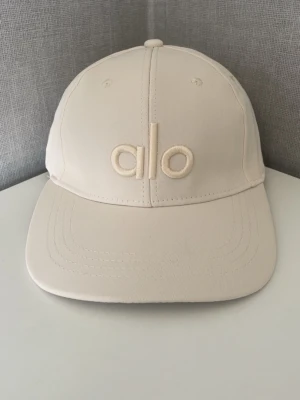 ALO keps i offwhite / Khaki - Snygg beige keps från Alo med broderad logga framtill. Klassisk böjd skärm och justerbar baksida. Tillverkad i ett mjukt och slitstarkt bomullstyg som passar perfekt till en avslappnad streetwear-look.