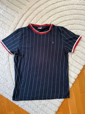 Mörkblå randig t-shirt med röd kant - Snygg mörkblå t-shirt med vita vertikala ränder och röda detaljer vid hals och ärmslut. Klassisk passform och mjukt material, perfekt för en avslappnad stil. Liten broderad logga på bröstet ger extra detalj. Superfin!