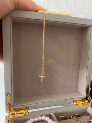 Guldhalsband med korshänge - Snyggt halsband i guld med ett litet korshänge dekorerat med gnistrande stenar. Kedjan är tunn och elegant, perfekt för att ge en stilren touch till din outfit. Korset har en klassisk form och stenarna ger extra bling. 