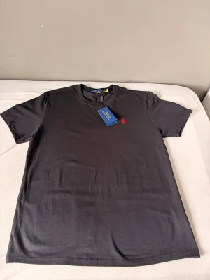 Svart t-shirt från Polo Ralph Lauren - Klassisk svart t-shirt från Polo Ralph Lauren med rund hals och den ikoniska röda logotypen broderad på bröstet. Tillverkad i mjuk bomull för en skön känsla och perfekt passform. Enkel och stilren design som funkar till allt.