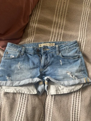 Blå denimshorts med slitningar - Säljer ett par ljusblå denimshorts från True Religion med slitna detaljer och uppvikta benslut. Klassisk femficksmodell med knapp och dragkedja. Perfekta för sommaren och ger en avslappnad streetstyle-look.